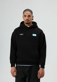 Sweat à capuche noir avec une poche avant, présentant des étiquettes de texte bleues "STRONGER" et "chance." Confectionné en matériau doux avec une coupe décontractée.