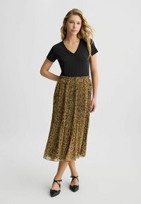 T-shirt nero a scollo a V abbinato a una gonna plissettata al ginocchio con stampa leopardata in tonalità di marrone e oro, indossata con ballerine nere appuntite.
