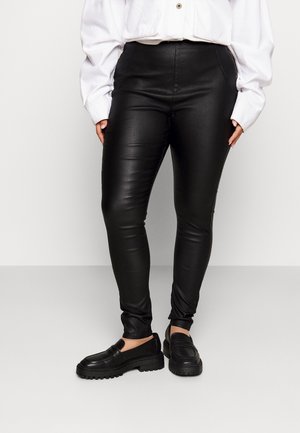 Persoon die zwarte skinny broek van imitatieleer, wit shirt met lange mouwen en zwarte loafers draagt, staand tegen een effen achtergrond.