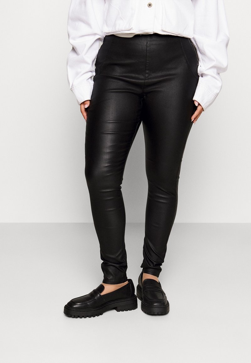 Personne portant un pantalon skinny simili cuir noir, un haut blanc à manches longues et des mocassins noirs, debout devant un fond uni.