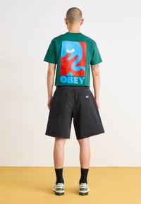 Green cotton t-shirt with a large red and blue graphic, featuring the word "OBEY." Paired with black denim shorts and sneakers.