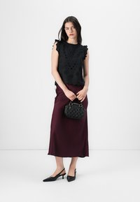 Črn vezen top z nabranimi rokavi, burgundska satenasta krila in majhna teksturirana črna torbica. Model nosi črne slingback petke.