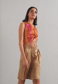 Satinový cropped top v oranžové a růžové barvě s uzlovým detailem, spárovaný s plisovanými béžovými šortkami, obě kousky mají hladké textury a uvolněný střih.