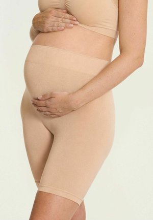 Zwangere persoon draagt beige zwangerschapsvormgevende shorts en top, en houdt voorzichtig haar buik vast met beide handen.