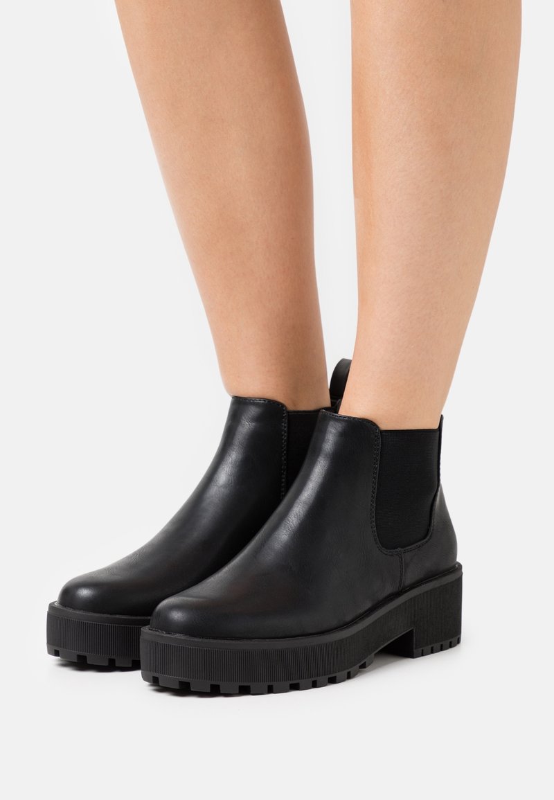 Bottes cheville en cuir noir avec semelles crantées épaisses portées sur des jambes nues sur un fond blanc uni.