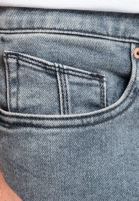 Jeans de mezclilla azul claro con un bolsillo trasero visible, que presentan costuras en contraste y un detalle de remache de cobre cerca de la cintura.