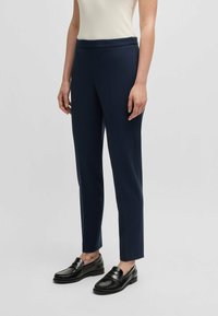 Pantaloni sartoriali blu navy con vestibilità slim, realizzati in un tessuto liscio. Presentano una zip laterale discreta e pences, abbinati a mocassini neri.