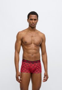 Κόκκινα ανδρικά εσώρουχα boxer με επαναλαμβανόμενο μοτίβο μαύρου λογότυπου «HUGO». Η μέση είναι μαύρη με κόκκινες λεπτομέρειες. Μαλακό, ελαστικό ύφασμα, μήκος μέχρι το μέσο του μηρού.