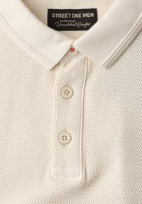 Gros plan d'un polo homme texturé crème avec col côtelé et trois boutons sur la patte de boutonnage.