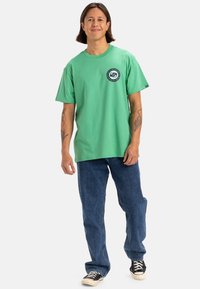 Camiseta verde de algodón con mangas cortas y un logo redondo en el pecho. Combinada con jeans de mezclilla azules y zapatillas negras.