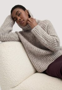 Kuscheliger Strickrollkragenpullover in Hellgrau, mit gerippter Textur und lässiger Passform. Getragen mit dunklen Hosen, sitzend auf einem weißen, flauschigen Sofa.