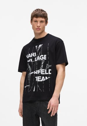 Ung mand iført en sort grafisk T-shirt med hvid forvrænget tekst og mørke mønstrede bukser, stående mod en ensfarvet hvid baggrund.