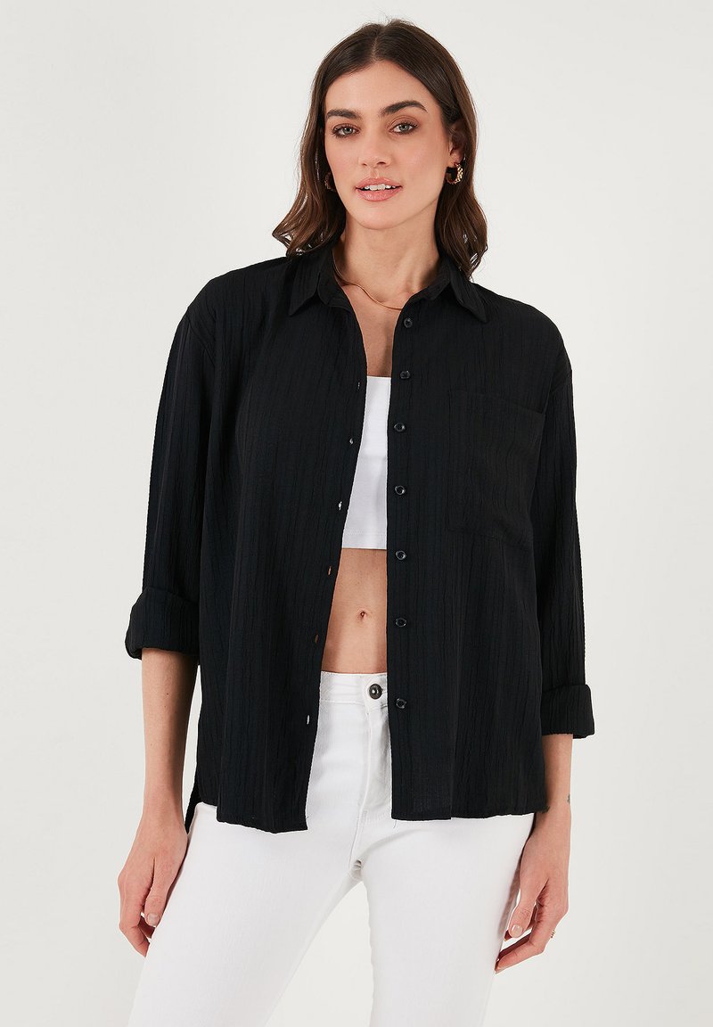 Black textured shirt with a button-up front, collar, and rolled sleeves, featuring a chest pocket, worn over a white top and white jeans.
