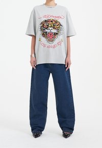 Šedé oversize tričko s barevným potiskem tygra a textem "Los Angeles", doplněné širokými tmav�ě modrými denimovými džínsy a špičatými černými botami.