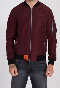 Giacca bomber bordeaux con zip frontale, polsini elasticizzati e tasca in vita. Presenta dettagli con etichetta arancione e decorazioni ricamate.