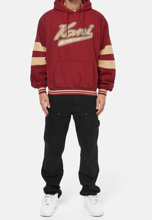 Vinröd huvtröja med guldfärgade bokstäver, med en framficka och randiga ärmar. Kombinerad med svarta cargobyxor och vita sneakers.