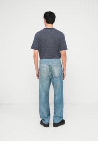 Άνδρας που στέκεται με την πλάτη σε λευκό τοίχο, φοράει μπλε και άσπρη ριγέ μπλούζα, ανοιχτόχρωμα τζιν και μαύρα παπούτσια.