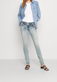 Light blue denim jeans with a slim fit and distressed detailing, paired with a white ribbed top and a light blue denim jacket. Black strap sandals.