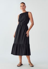 Black sleeveless dress with a gathered neckline and an elastic waist featuring a silver buckle, ending in a ruffled hem. Smooth fabric texture.