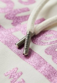 Tejido de sudadera blanca con un gráfico de purpurina rosa brillante. Presenta un cordón beige con aglets plateados, grabados con "SUPERDRY."
