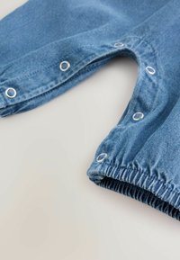 Pantalon bébé en jean bleu avec boutons-pression le long des jambes intérieures et poignets élastiques sur fond blanc.