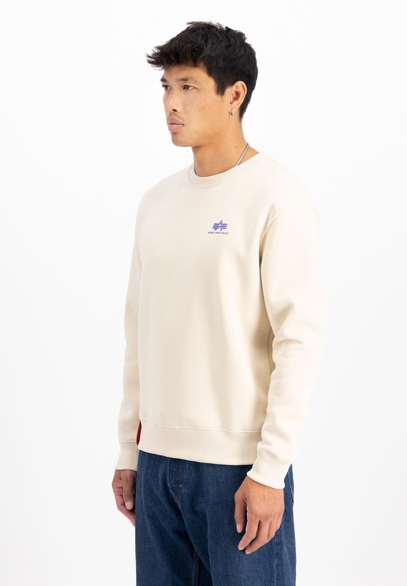 Beige sweatshirt med ribbet rund halsudskæring, lilla logo på brystet og rød detalje i kanten. Stoffet har en glat, bomuldsagtig tekstur.