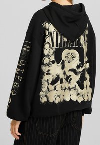 Sudadera negra oversize con un estampado gráfico dorado de flores y patrones circulares en la espalda y el texto "IN UTERO" en la manga.