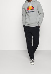 Sudadera con capucha gris con un gran logo gráfico rojo y amarillo en el frente, combinada con pantalones negros y zapatillas negras. Ajuste casual y textura suave.