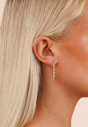 Guldtonade örhängen med fem sammanlänkade hängen av kristaller i klara och gula nyanser, mot en texturerad bakgrund.