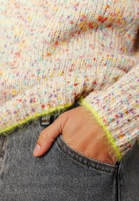 Pull en maille crème pâle avec des taches multicolores, doté d'un ourlet jaune duveteux. La main repose dans une poche en denim gris.