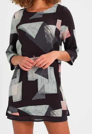 Vrouw die een zwarte jurk draagt met abstracte geometrische vormen in grijs, roze en wit, met driekwartmouwen en een ronde hals.