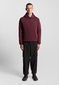 Bordeaux hoodie med høj krave, parret med lette sorte cargobukser og mørke træningssko, står op ad en neutral baggrund.