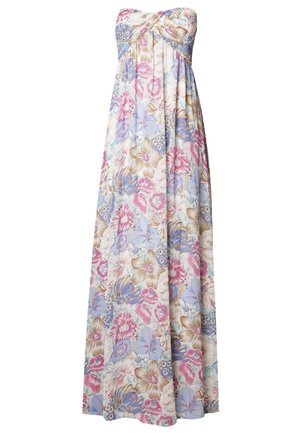Personne portant une veste en jean foncée sur une robe maxi fleurie colorée, tenant un chapeau beige à côté.