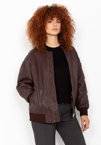 Veste bombardier en cuir marron avec un design matelassé, des poignets côtes et une fermeture éclair. Modèle la portant avec une chemise noire et un pantalon gris.