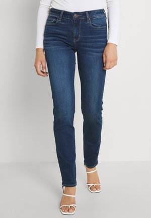 Vrouw draagt strakke donkerblauwe spijkerbroek, wit shirt met lange mouwen en witte open-teen sandalen met hak, staand tegen een effen achtergrond.