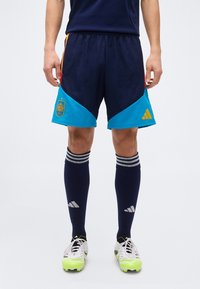 Male athlete wearing navy and turquoise Spain soccer shorts, navy knee-high socks with white stripes, and white cleats with neon yellow accents.