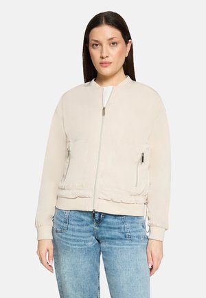 Kvinde med glat mørkt hår iført en lys beige bomberjakke med lynlås og blå jeans, stående foran en ensfarvet baggrund.