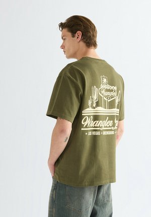 Młody mężczyzna o krótkich brązowych włosach, noszący oliwkowo-zielony t-shirt Wrangler z motywem kaktusa i Las Vegas na plecach.