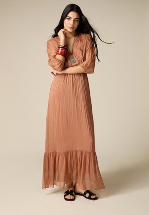 Donna con un lungo abito marrone chiaro con maniche 3/4, che indossa sandali neri, braccialetti colorati e una grande collana con pendente, in piedi con le braccia incrociate.