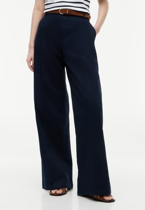 Femme portant un pantalon large bleu marine, ceinture marron, haut rayé blanc et bleu marine, et sandales ouvertes marron, debout les mains dans les poches.