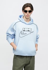 Lichtblauwe sweatvest met capuchon, zwart logodetail, kangoeroezak en contrasterende naden. Draagt groene cargobroek en smalle zonnebril.