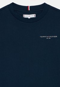 Tamsiai mėlynas medvilninis marškinėlis su apvalia kaklo linija, ant kurio yra mažas baltas logotipas ir raudonai baltos juostelės detalė kaklo liniuotėje.