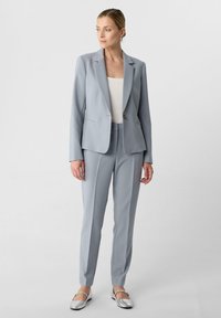 Completo grigio chiaro su misura con giacca aderente e pantaloni slim, realizzato in tessuto morbido. Dettagli notevoli includono un solo bottone e design minimalista.