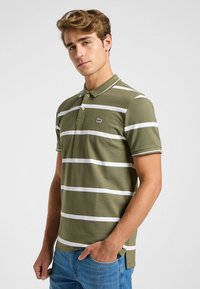 Polo en verde oliva con rayas horizontales blancas, tapeta de dos botones y pequeño logotipo en el pecho. Textura suave y mangas cortas.