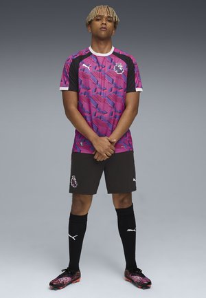 Puma futbola tērps, kas sastāv no rozā un violetā raksta krekla, melniem šortiem un atbilstošām zeķēm ar baltām akcentiem un lauvas logotipu.