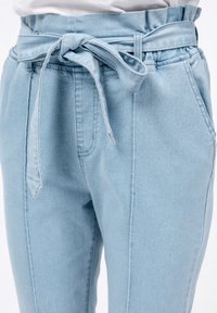 Pantaloni in denim blu chiaro presentano una vita ampia con un nodo, tasche laterali e cuciture evidenti lungo le gambe.