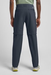 Pantaloni convertibili grigio scuro con una texture liscia, tasca posteriore con zip e orli regolabili. Abbinati a scarpe da trekking di colore chiaro.
