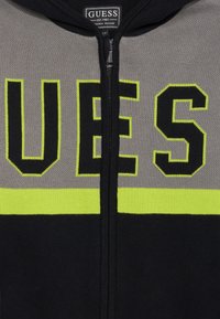 Hoodie zippé noir et gris avec des accents jaune fluo et le texte "GUESS" affiché de manière proéminente. Tissu tricoté avec une texture lisse.