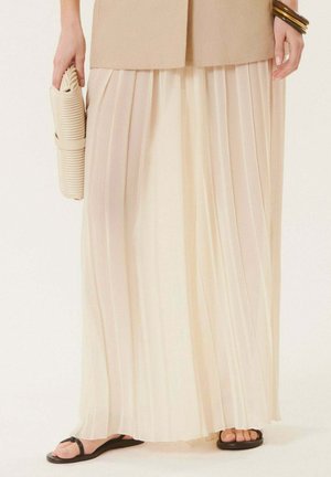 Jupe longue plissée beige portée avec des sandales plates noires et un blazer beige, tenant une pochette beige texturée d'une main.