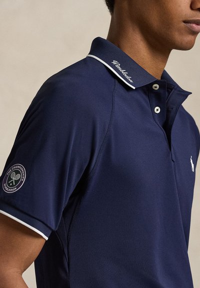 Άτομο που φοράει μπλε κονκάρδα πόλο του Wimbledon με λευκό περίγραμμα, κεντημένο λογότυπο του Wimbledon στο μανίκι και μικρό λευκό έμβλημα αλόγου στο στήθος.